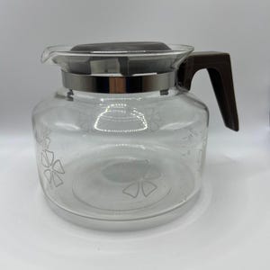 May include: A clear glass coffee carafe featuring a dark brown plastic handle and a matching lid. The glass body is decorated with a white floral pattern and includes printed measurement markings on the side, indicating up to 10 cups of capacity.