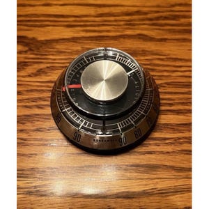 May include: A round Honeywell thermostat with a brushed silver metal outer ring and a black inner dial. The device features black numerical markings from 60 to 90, a central silver circular knob, and a thin red indicator line on the black face.