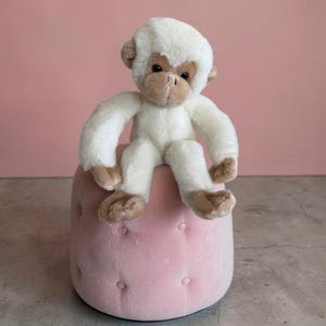 Pode incluir: Um brinquedo de pelúcia em forma de macaco com pelo branco macio no corpo e na cabeça, apresentando detalhes em castanho claro no rosto, orelhas, mãos e pés. O macaco tem olhos escuros e um nariz pequeno, sentado com os braços e pernas para a frente.