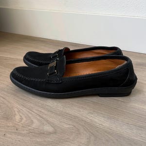 Puede incluir: Un par de mocasines negros de ante con hebilla dorada. Los zapatos tienen la punta redondeada y un dise&ntilde;o cosido en la parte superior. El interior es de color marr&oacute;n claro y las suelas son negras.