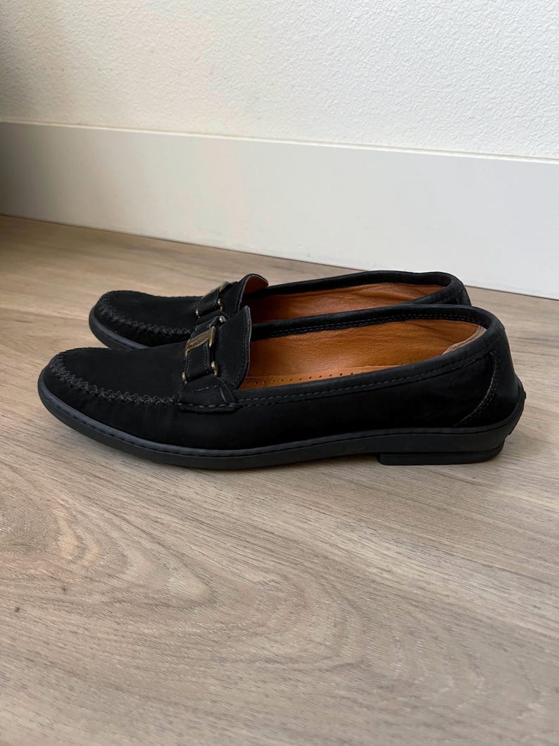 Puede incluir: Un par de mocasines negros de ante con hebilla dorada. Los zapatos tienen la punta redondeada y un dise&ntilde;o cosido en la parte superior. El interior es de color marr&oacute;n claro y las suelas son negras.