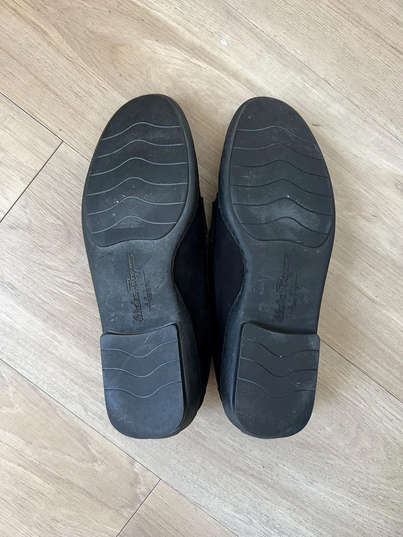 Puede incluir: Un par de mocasines de cuero negro, suelas hacia arriba, con el nombre de la marca "Salvatore Ferragamo" grabado en la suela. Las suelas tienen un dise&ntilde;o texturizado y muestran signos de desgaste.