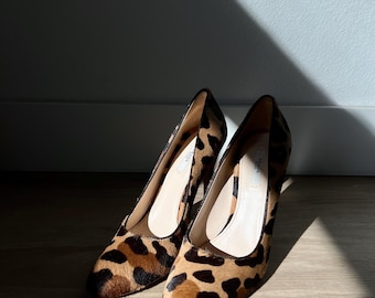 Puede incluir: Un par de tacones Prada con estampado de leopardo. Los zapatos tienen punta afilada y tacón medio. El interior del zapato es de color beige claro. Los zapatos están sobre un suelo de madera.