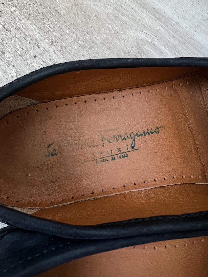 Puede incluir: Primer plano del interior de un zapato negro. La plantilla del zapato es de cuero marr&oacute;n con las palabras "Salvatore Ferragamo SPORT MADE IN ITALY" impresas en verde.