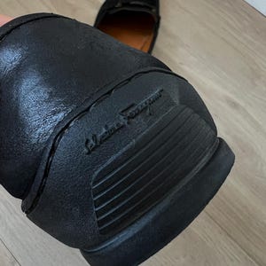 Puede incluir: Mocas&iacute;n de cuero negro con suela de goma. La suela presenta el nombre de la marca en relieve. El zapato tiene un dise&ntilde;o cl&aacute;sico, adecuado para diversas ocasiones. El segundo zapato es visible en el fondo.