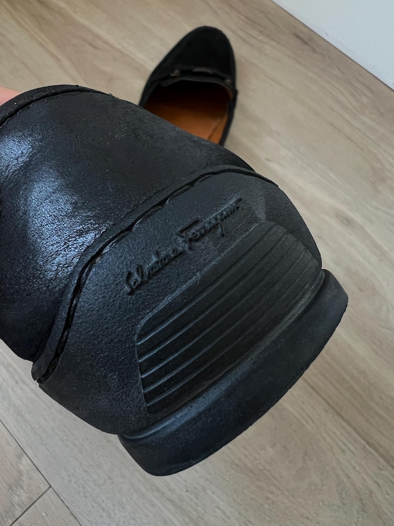 Puede incluir: Mocas&iacute;n de cuero negro con suela de goma. La suela presenta el nombre de la marca en relieve. El zapato tiene un dise&ntilde;o cl&aacute;sico, adecuado para diversas ocasiones. El segundo zapato es visible en el fondo.