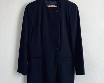 Puede incluir: Un blazer azul marino con cierre de un solo botón y dos bolsillos delanteros. El blazer está colgado de una percha de madera. El nombre de la marca "ESCADA" es visible en el cuello.