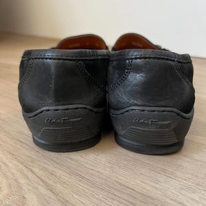 Puede incluir: Un par de mocasines de cuero negro con tac&oacute;n bajo. Las suelas son de color gris oscuro y llevan el nombre de la marca. Los zapatos est&aacute;n sobre una superficie de madera clara.