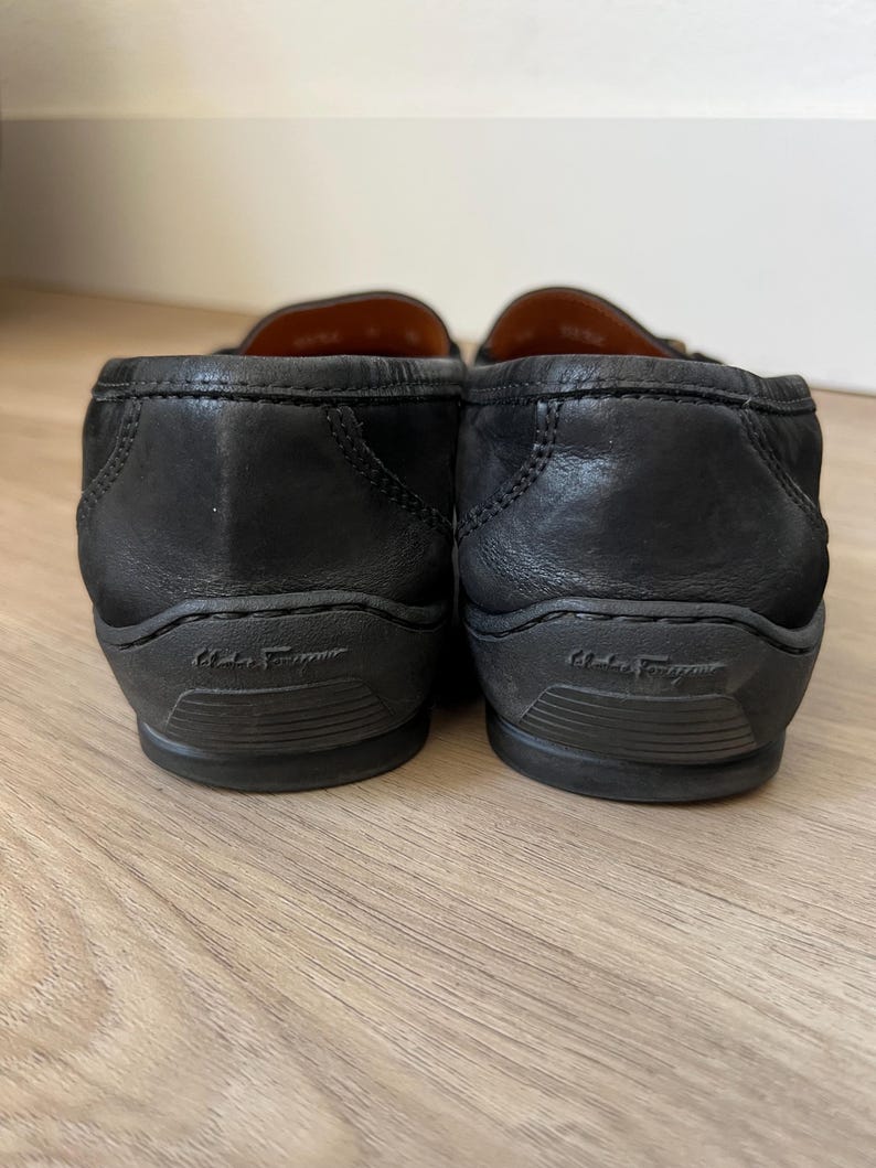 Puede incluir: Un par de mocasines de cuero negro con tac&oacute;n bajo. Las suelas son de color gris oscuro y llevan el nombre de la marca. Los zapatos est&aacute;n sobre una superficie de madera clara.