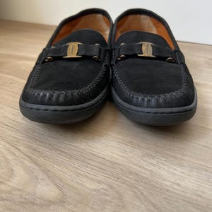 Puede incluir: Un par de mocasines negros de ante con un dise&ntilde;o cosido alrededor de la puntera y una hebilla dorada. El forro interior es de color naranja c&aacute;lido. Estos zapatos son de estilo cl&aacute;sico.