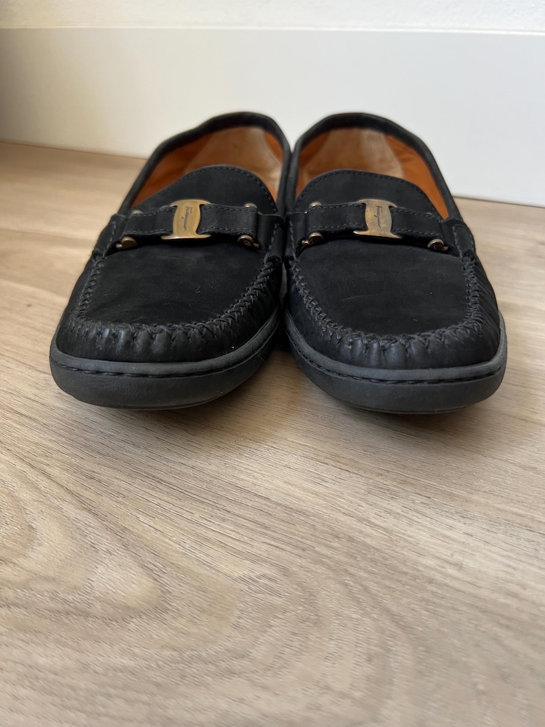 Puede incluir: Un par de mocasines negros de ante con un dise&ntilde;o cosido alrededor de la puntera y una hebilla dorada. El forro interior es de color naranja c&aacute;lido. Estos zapatos son de estilo cl&aacute;sico.