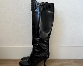 Puede incluir: Botas negras hasta la rodilla con acabado brillante. Las botas tienen punta y tacón de aguja. La parte superior de la bota presenta un patrón tejido decorativo y un detalle de hebilla. Las botas están hechas de un material brillante.
