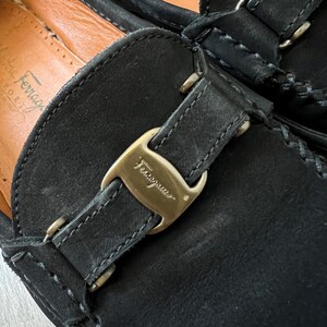 Puede incluir: Primer plano de mocasines de cuero negro con hebilla dorada. Los mocasines tienen costuras visibles y un interior de cuero. La marca "Ferragamo" es visible en la hebilla.
