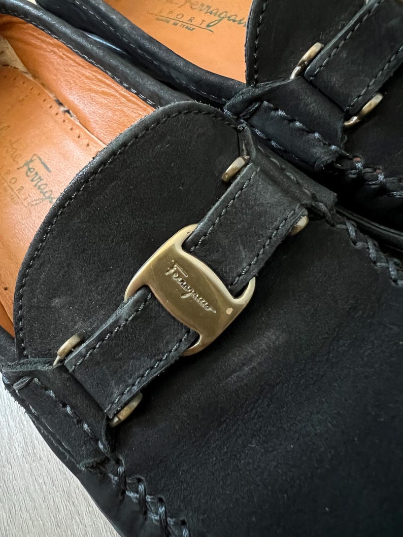 Puede incluir: Primer plano de mocasines de cuero negro con hebilla dorada. Los mocasines tienen costuras visibles y un interior de cuero. La marca "Ferragamo" es visible en la hebilla.