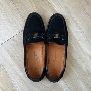 Puede incluir: Un par de mocasines de cuero negro con hebilla dorada. Los zapatos tienen un dise&ntilde;o cl&aacute;sico con punta redonda y empeine cosido. El interior del zapato es de color marr&oacute;n claro. Los zapatos est&aacute;n sobre una superficie de madera clara.