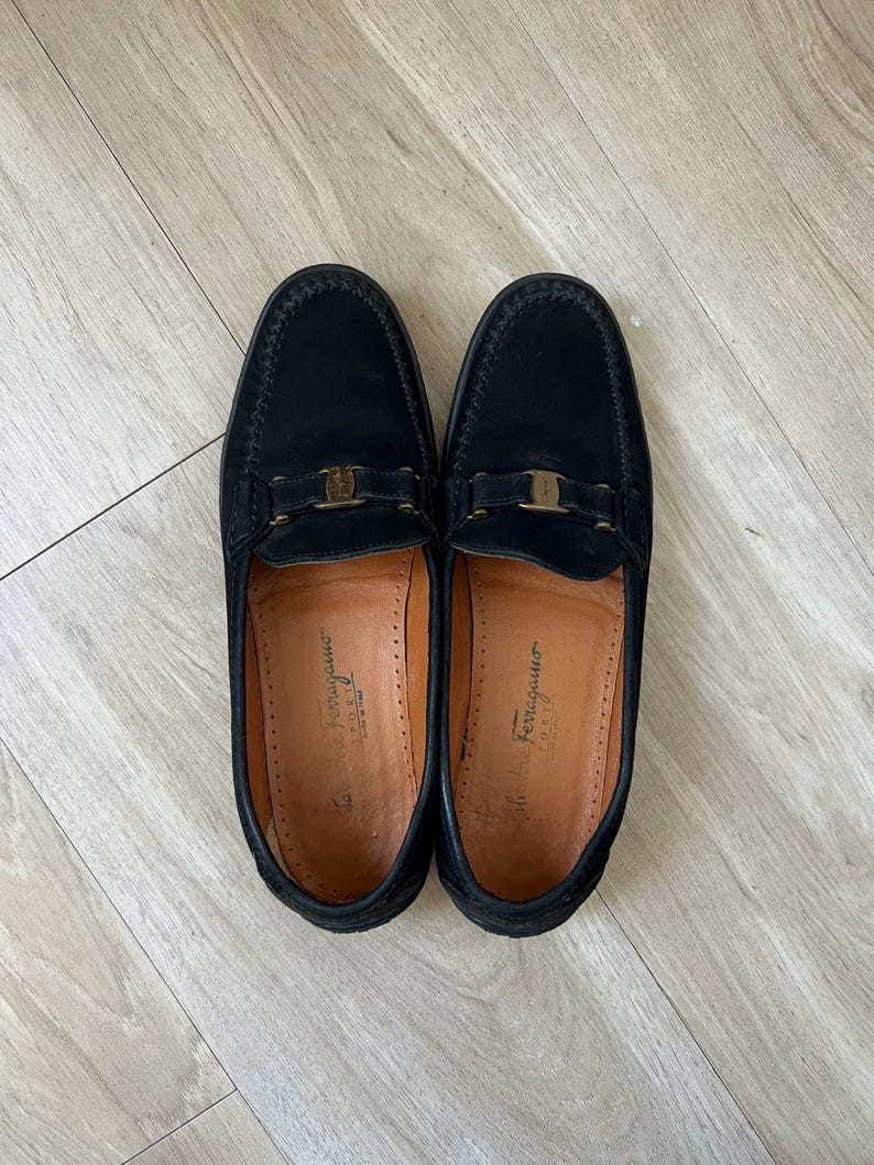 Puede incluir: Un par de mocasines de cuero negro con hebilla dorada. Los zapatos tienen un dise&ntilde;o cl&aacute;sico con punta redonda y empeine cosido. El interior del zapato es de color marr&oacute;n claro. Los zapatos est&aacute;n sobre una superficie de madera clara.