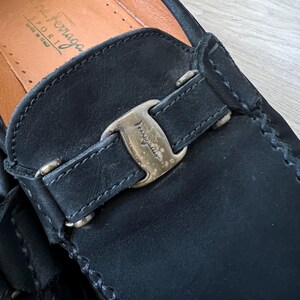 Puede incluir: Primer plano de un mocas&iacute;n de cuero negro con hebilla dorada. El zapato presenta costuras visibles y una plantilla de cuero. La marca "Salvatore Ferragamo" es visible en la plantilla.
