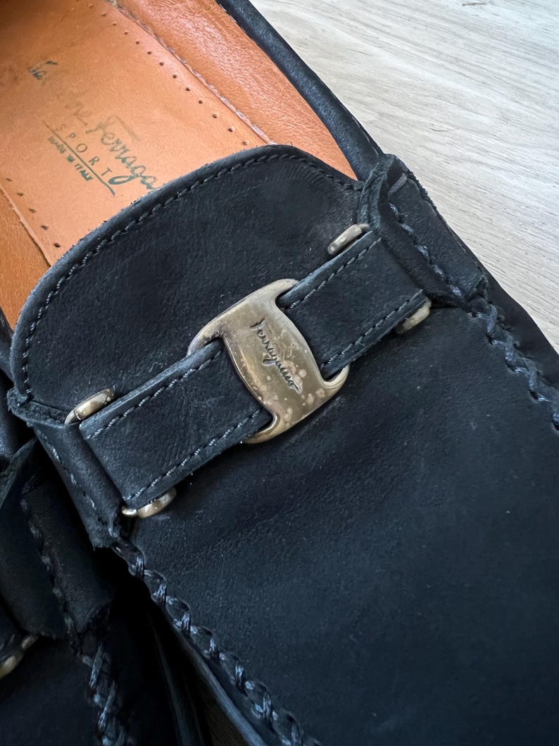 Puede incluir: Primer plano de un mocas&iacute;n de cuero negro con hebilla dorada. El zapato presenta costuras visibles y una plantilla de cuero. La marca "Salvatore Ferragamo" es visible en la plantilla.