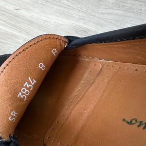Puede incluir: Primer plano del interior de un zapato de cuero negro. El interior del zapato es de cuero marr&oacute;n, con la talla 8 y el n&uacute;mero 3834 impresos en blanco. Tambi&eacute;n es visible el nombre de la marca del zapato.