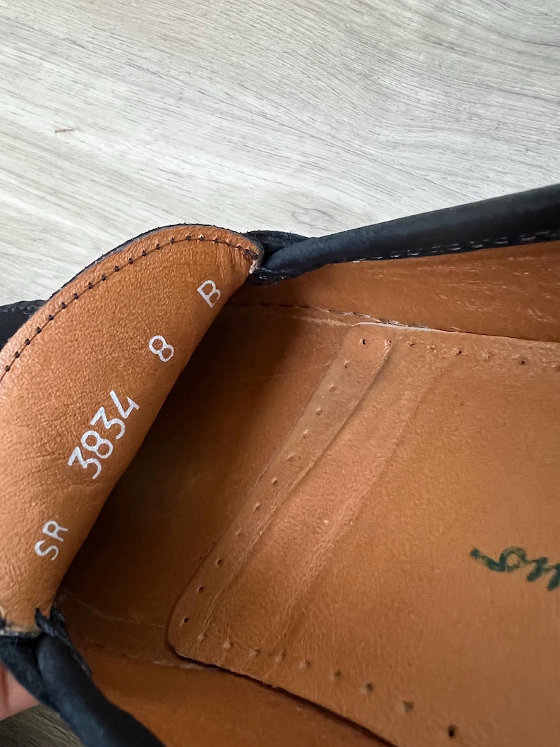 Puede incluir: Primer plano del interior de un zapato de cuero negro. El interior del zapato es de cuero marr&oacute;n, con la talla 8 y el n&uacute;mero 3834 impresos en blanco. Tambi&eacute;n es visible el nombre de la marca del zapato.