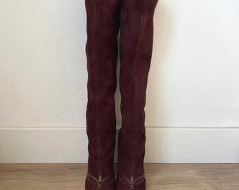 Puede incluir: Un par de botas altas de ante burdeos. Las botas tienen puntera afilada con un acento marrón oscuro brillante y un detalle decorativo de lazo. Las botas están de pie contra una pared blanca.