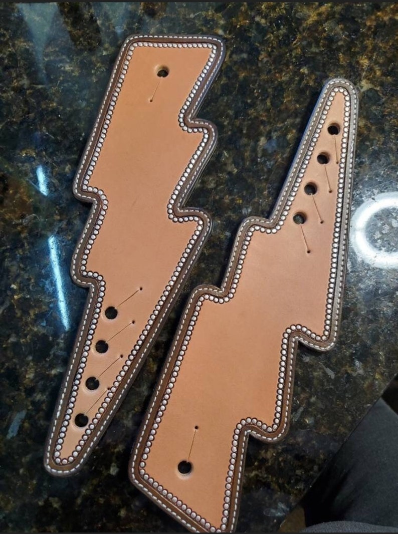 May include: Two brown leather lightning bolt shapes with white stitching and silver studs around the edges. The leather is a light brown colour and the stitching is a darker brown colour.