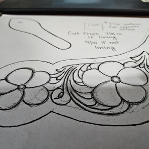 May include: A pencil sketch of a leather craft design, featuring floral and scrollwork patterns. The design includes two large flowers and a separate strap pattern. Text indicates cutting instructions for the leather.