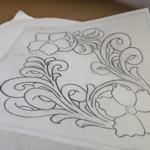 May include: A detailed pencil drawing of a floral design on white paper. The design features two large flowers and intricate swirling leaf and vine patterns. The drawing is in a corner layout, with the design filling the space.