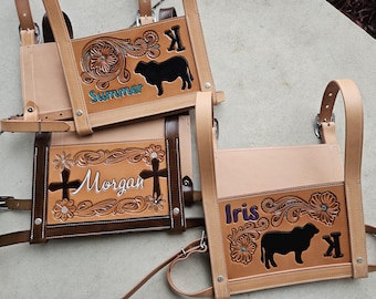 May include: Three brown leather bags with tooled designs and personalized names. The bags have a strap and a buckle. The names on the bags are "Summer", "Morgan", and "Iris".