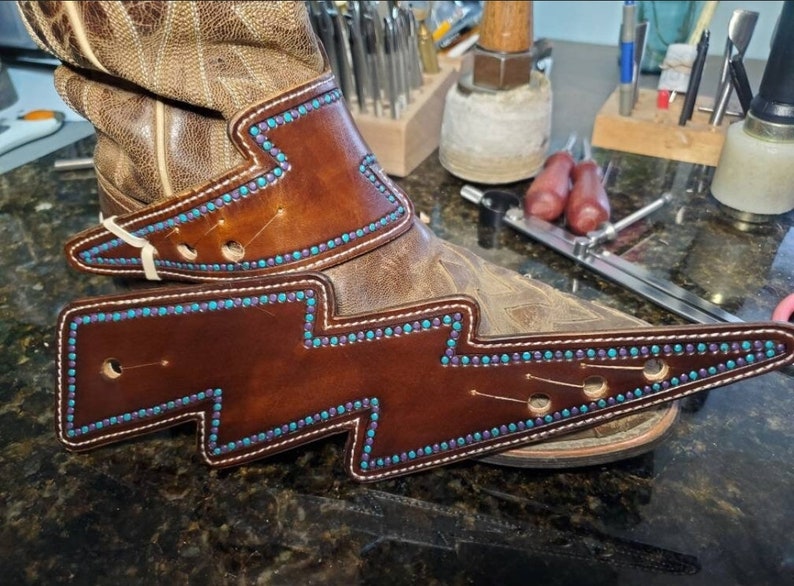 May include: Brown leather boot spur straps with turquoise and purple stitching and accents. The straps are decorated with a lightning bolt design.