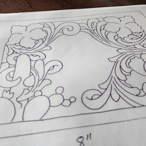 May include: A black and white floral and cactus design on a white paper. The design is 8 inches by 6 inches. The design features a central open space surrounded by detailed floral and cactus motifs, with swirling leaves and flower shapes.