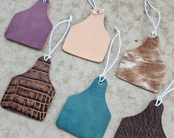 May include: A collection of six leather cow tags in various colors and textures. The tags include a purple, tan, brown crocodile print, teal, brown, and a cowhide pattern. Each tag has a white string attached for hanging.