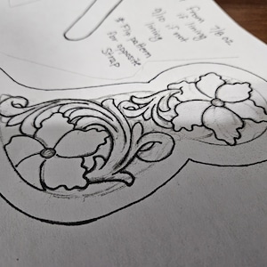 May include: A detailed pencil sketch of a leather craft design, featuring floral and scrollwork patterns. The design includes two large flowers and intricate leaf details, outlined in black on a white background. The sketch is for a strap.