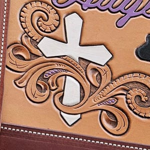 May include: Close-up of a tan leather item with a white cross and decorative scrollwork. The cross is centered, with the scrollwork and purple text above. The item has a brown border and a silver button.