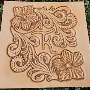 Peut inclure: Une pièce de cuir carrée, brun clair, avec un motif floral et la lettre "K" gravée. Le motif présente deux grandes fleurs et des feuilles tourbillonnantes. Le cuir est de couleur naturelle et la gravure crée un effet 3D.