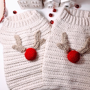 Könnte beinhalten: Zwei cremefarbene, gehäkelte Hundepullover mit Rentiergeweih und roter Nase. Die Pullover haben einen gerippten Kragen und sind mit beigefarbenem Geweih und einer roten Pompon-Nase verziert, ideal für die Weihnachtszeit.