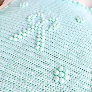 Könnte beinhalten: Eine hellgrüne gehäkelte Babydecke mit einem erhabenen Muster aus weißen Pompons.