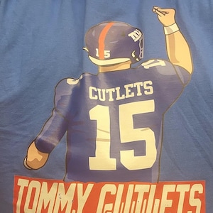 May include: Blue t-shirt with a cartoon image of a football player wearing a blue jersey with the number 15 and the text "CUTLETS". The text "TOMMY CUTLETS" is printed in red and white on the bottom of the shirt.