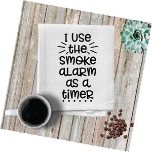 May include: White kitchen towel with black text that reads "I use the smoke alarm as a timer..." The towel is on a wood background with coffee beans and a succulent plant.