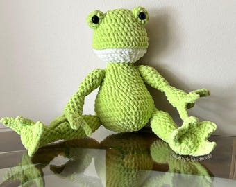 May include: A handmade, light green crochet frog with black eyes and a white collar. The frog is sitting with its legs spread out. The frog's feet and hands are detailed with three toes. The frog is made of a soft, textured yarn.