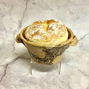 May include: A round loaf of bread dusted with flour sits in a rustic, ceramic bowl. The bowl has a tan and brown marbled design with two handles. The bread is golden brown with a scored top.