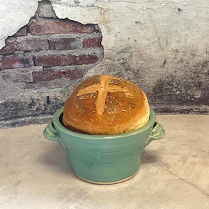 May include: A round loaf of bread with a cross-shaped score, speckled with herbs, sits in a turquoise ceramic bowl with handles. The background features a distressed brick and plaster wall.
