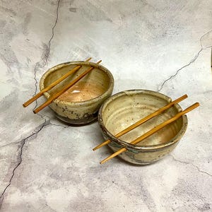 May include: Two handmade ceramic bowls with a speckled beige and brown glaze. Each bowl has a pair of wooden chopsticks resting across the rim. The bowls are designed for serving Asian cuisine.