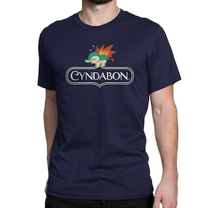 May include: A navy blue t-shirt featuring a white graphic of a cartoon water-type Pokemon with a flame on its back. The graphic is surrounded by a white oval with the text "CYNDABON" in white.