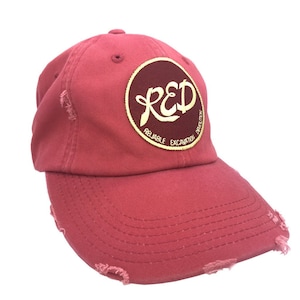 Könnte beinhalten: Verwaschene rote Baseballkappe mit gebogenem Schirm. Auf der Kappe befindet sich ein kreisförmiger Aufnäher mit dem Wort "RED" in einer stilisierten Schrift, umgeben von den Wörtern "Reliable Excavation" und "Disruption".