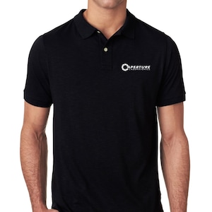 Peut inclure: Un polo noir avec le logo d'Aperture Laboratories sur la poitrine gauche. Le logo est un cercle blanc avec un symbole d'ouverture blanc à l'intérieur et le texte "Aperture Laboratories" en dessous.