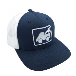 May include: Navy blue trucker cap with a white mesh back and a curved peak. The front displays a rectangular patch with a white tow truck graphic within a navy blue border. The cap has a button on top.