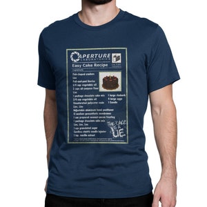 Op de afbeelding: Een marineblauw t-shirt met een grafische print van een Aperture Laboratories Easy Cake recept. Het ontwerp bevat een ingrediëntenlijst, een kleine afbeelding van een chocoladetaart met een kaarsje en de tekst "THE CAKE IS A LIE".