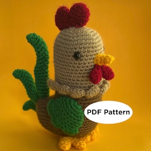Op de afbeelding: Gehaakt amigurumi speelgoed in de vorm van een haan met een rode kam, groene staartveren, een bruin lichaam en gele voeten. Het speelgoed zit op een gele achtergrond. PDF-patroon.