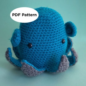 Puede incluir: Un juguete de peluche de pulpo tejido a crochet con un cuerpo azul y tentáculos grises. El pulpo tiene ojos negros y una expresión amigable. El texto "PDF Pattern" es visible en la imagen.